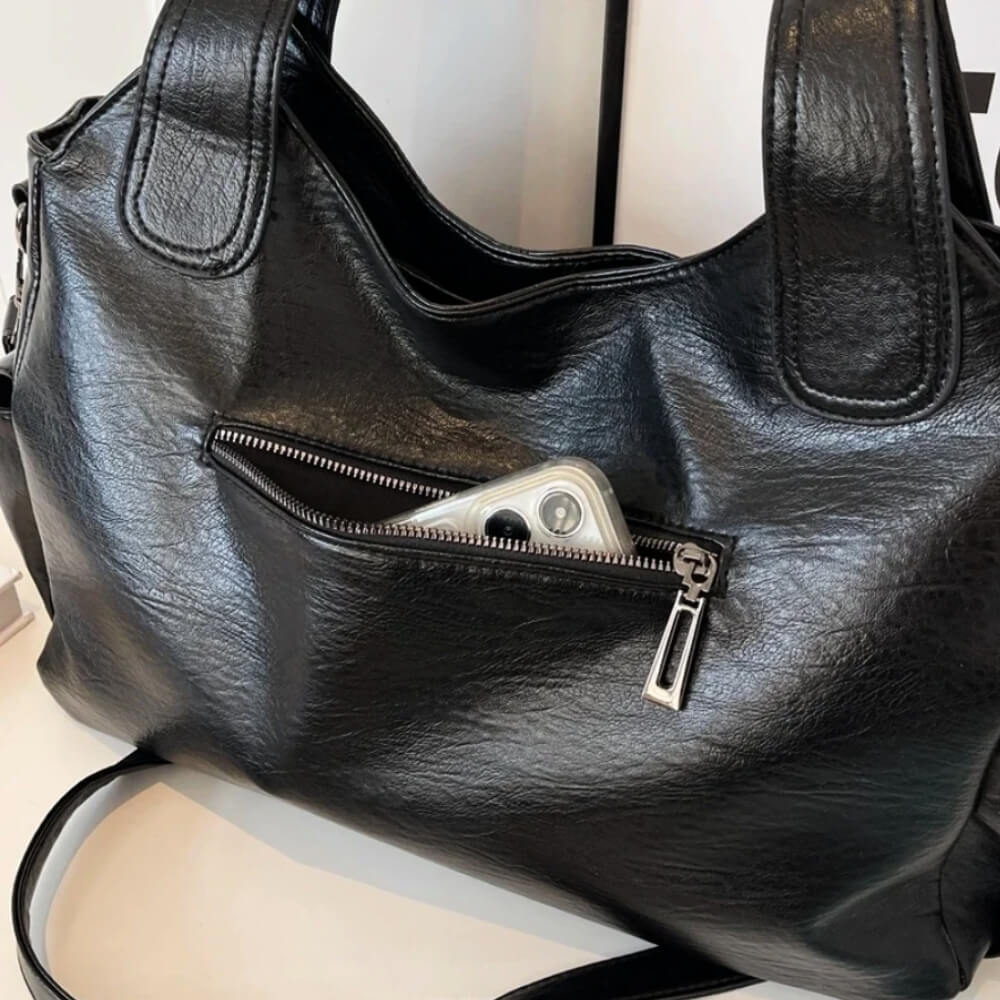 Schwarze Lederhandtasche mit seitlichem Reißverschlussfach, in dem ein Smartphone steckt.