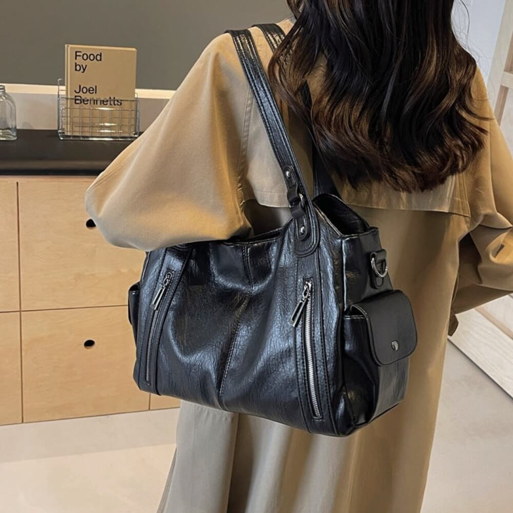 Frau mit beigem Mantel trägt eine schwarze Lederhandtasche mit mehreren Fächern über der Schulter.