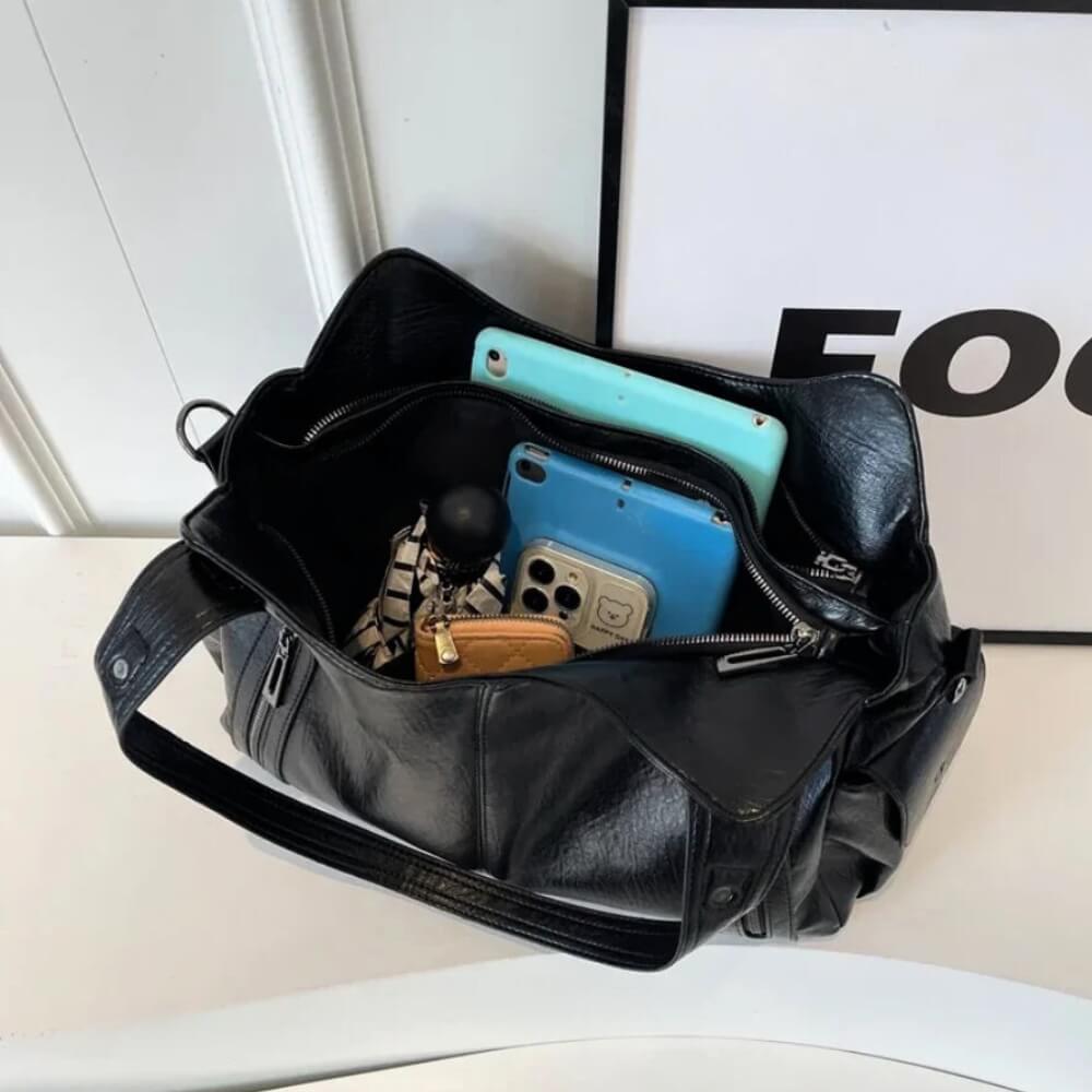 Geöffnete schwarze Handtasche mit geräumigem Innenraum, gefüllt mit Tablet, Smartphone, Portemonnaie und weiteren Alltagsgegenständen.