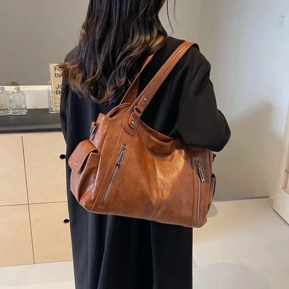 Frau in schwarzem Mantel trägt eine cognacfarbene Lederhandtasche mit seitlichen Taschen über der Schulter.